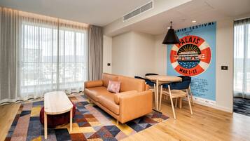 A modern hotel room with a sofa, a small table, and a chair. There is a large window with sheer curtains, a patterned rug, and a wall with a poster advertising an event.