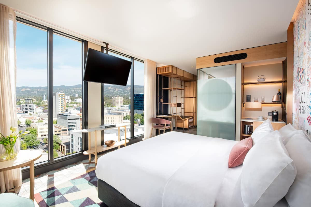 A modern hotel room with a large bed, a TV, a desk, and a city view.
