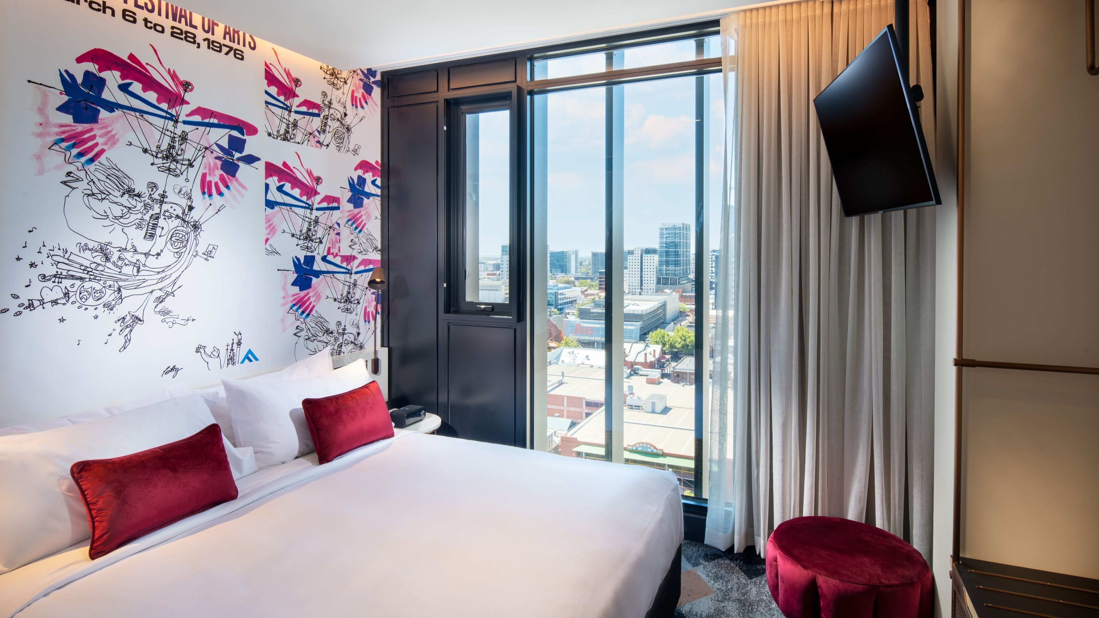 A hotel room with a large bed, a city view, a flat-screen TV, and a wall mural.