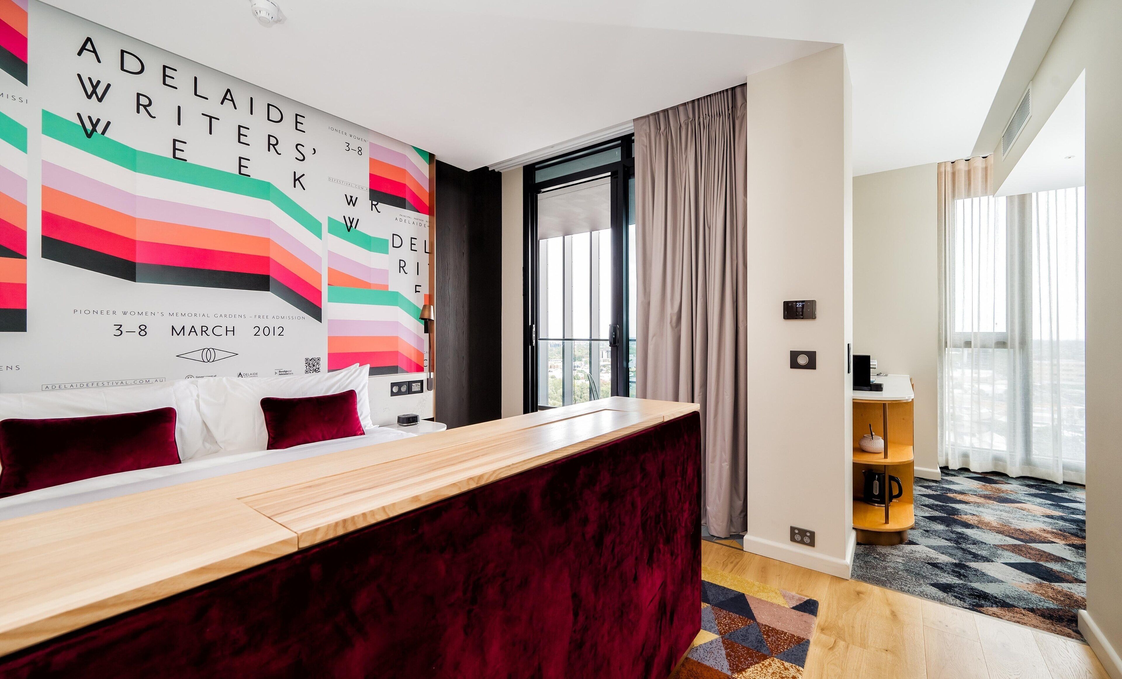 A modern hotel room with a wooden desk, a red velvet headboard, and a large poster on the wall.