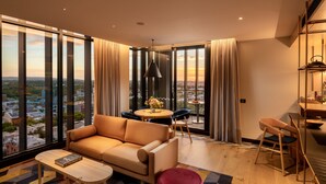 A modern living room with a sofa, coffee table, and ottoman, a dining area with a table and chairs, and large windows offering a city view. - Hotel Indigo Adelaide Markets by IHG (Adelaide)