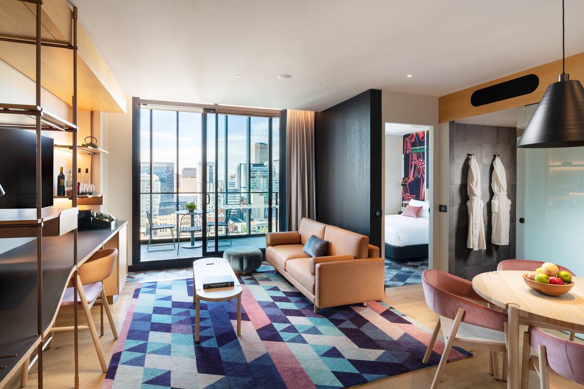 A modern hotel room with a large window, a sofa, a dining table with chairs, a TV, and a minibar.