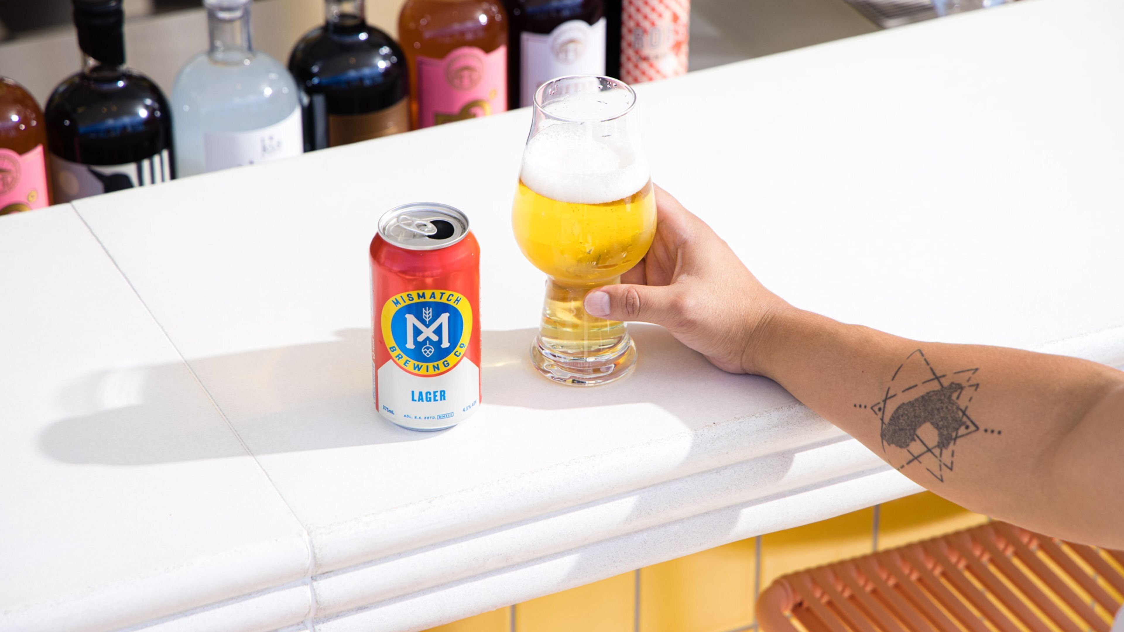 Una mano sosteniendo una jarra de cerveza junto a una lata de cerveza sobre una encimera con varias botellas al fondo.