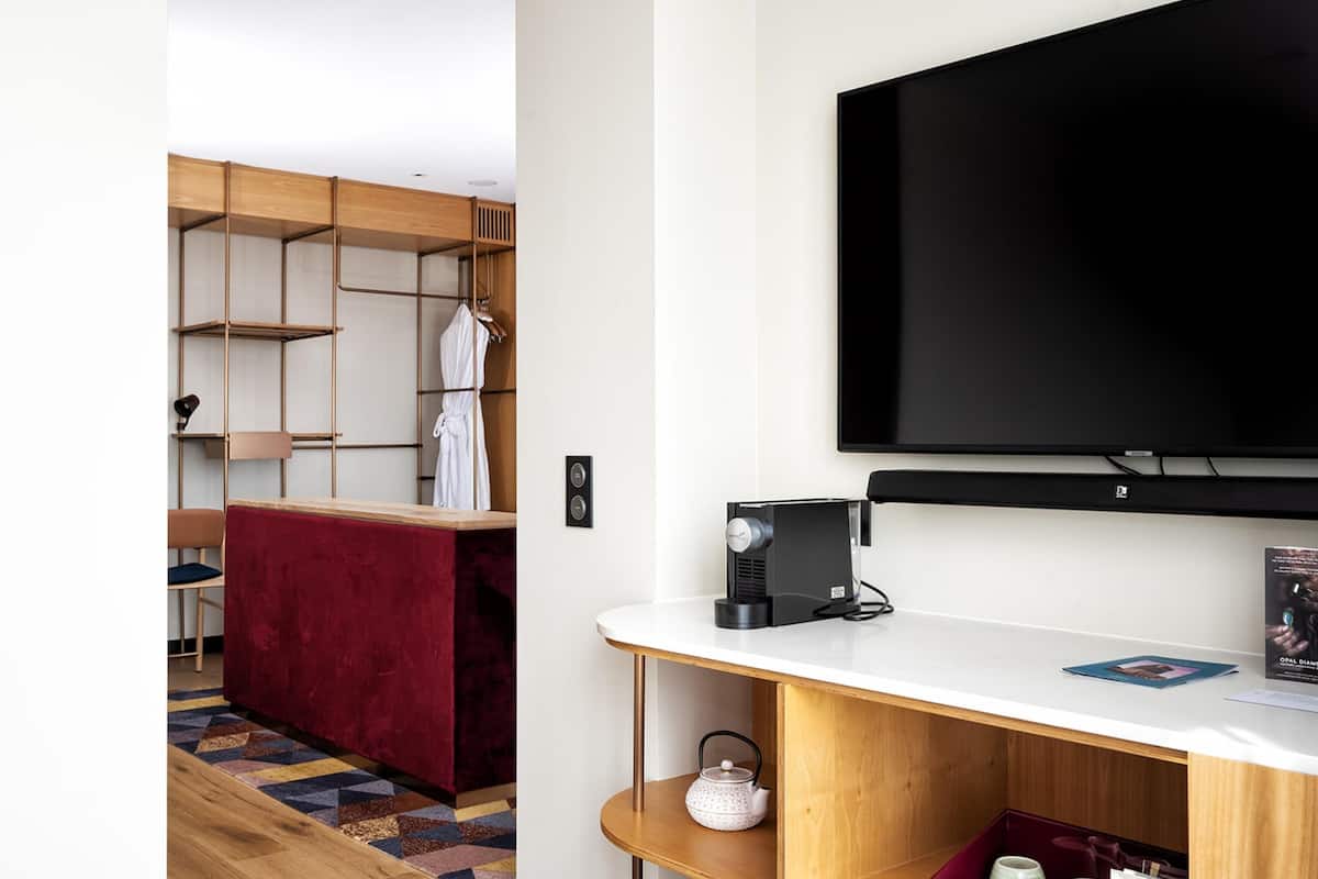 A modern hotel room with a large flat-screen TV, a wooden desk, a red chair, a patterned rug, and a wardrobe with multiple shelves.