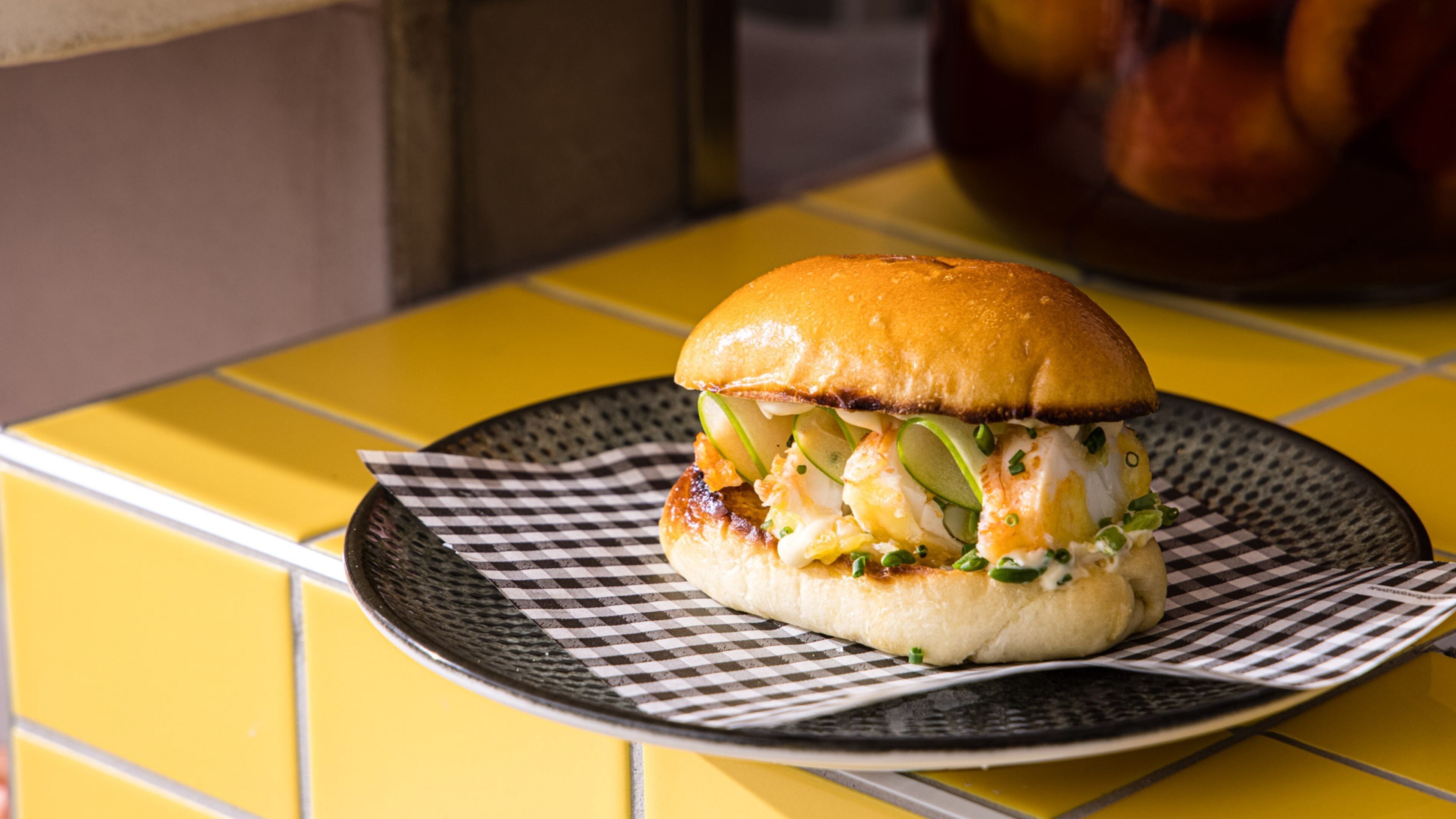 A sandwich on a checkered paper on a yellow table.