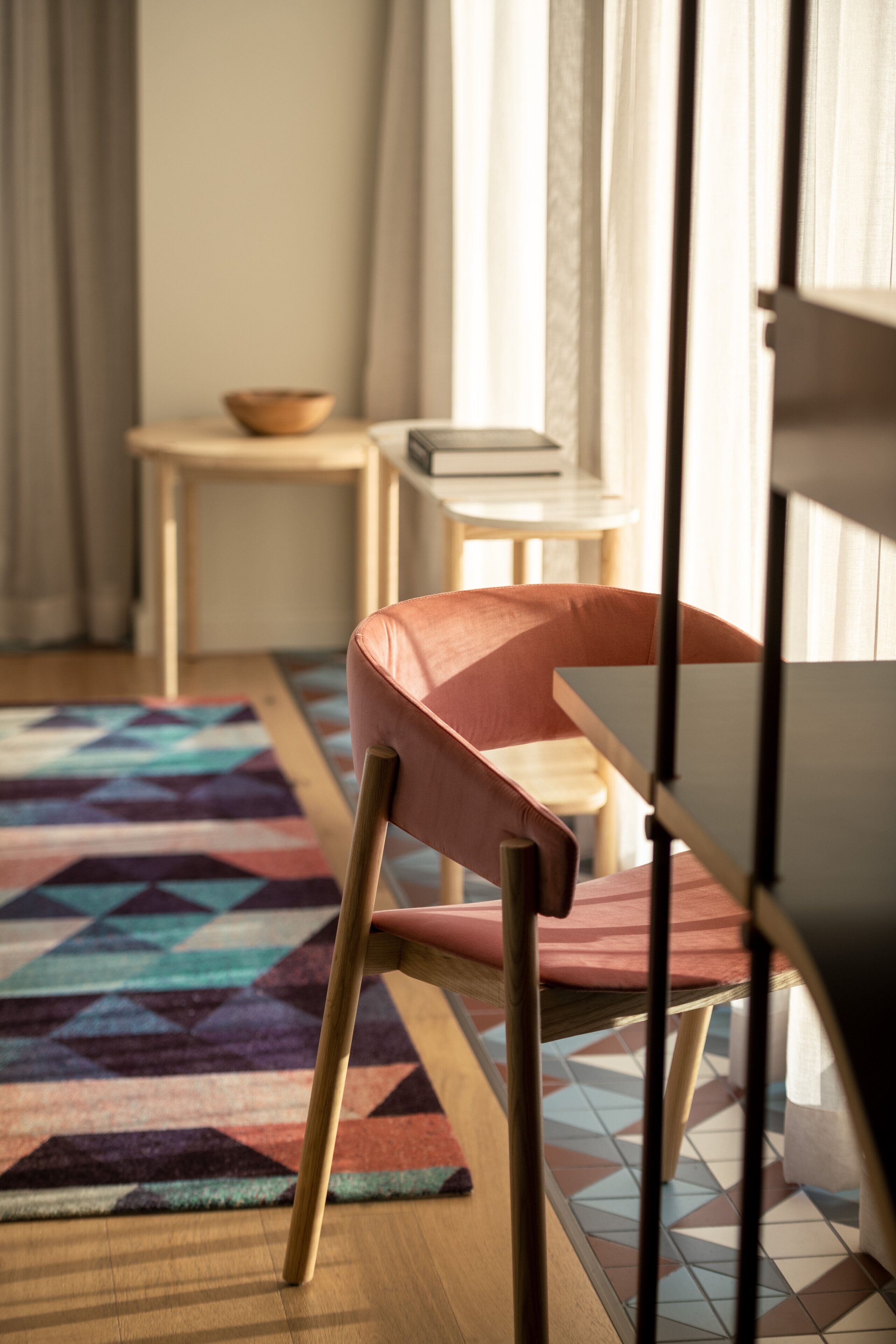 A modern room with a wooden floor, a patterned rug, a pink chair, a small table with a bowl, and a window with curtains.