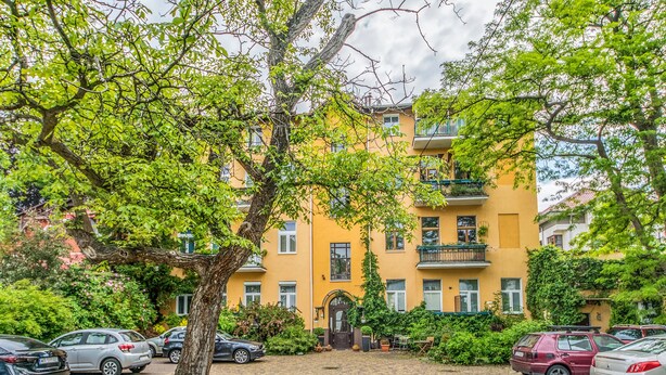 A yellow building with a central entrance, surrounded by parked cars and trees. - Victus Apartamenty - Meru (Sopot)