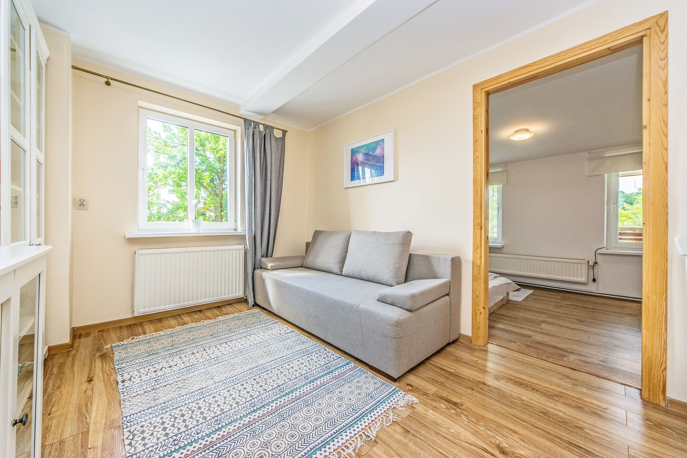 A room with a grey sofa, a patterned rug, wooden flooring, a window with curtains, and a painting on the wall.