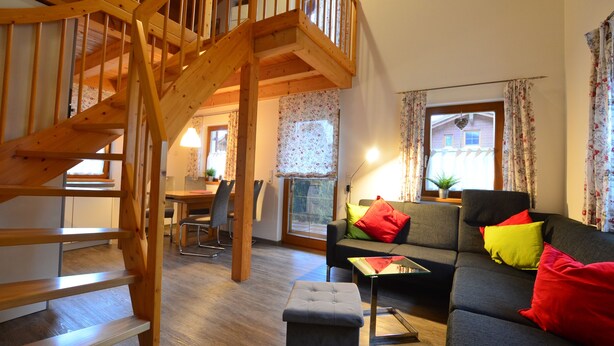 Een houten trap leidt naar een verdieping, een woonkamer met een bank en kleurrijke kussens, een eethoek met stoelen en een tafel, en een raam met gordijnen.