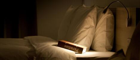 Una habitación de hotel tenuemente iluminada con una cama, una lámpara de noche y una mesita de madera con un libro titulado
