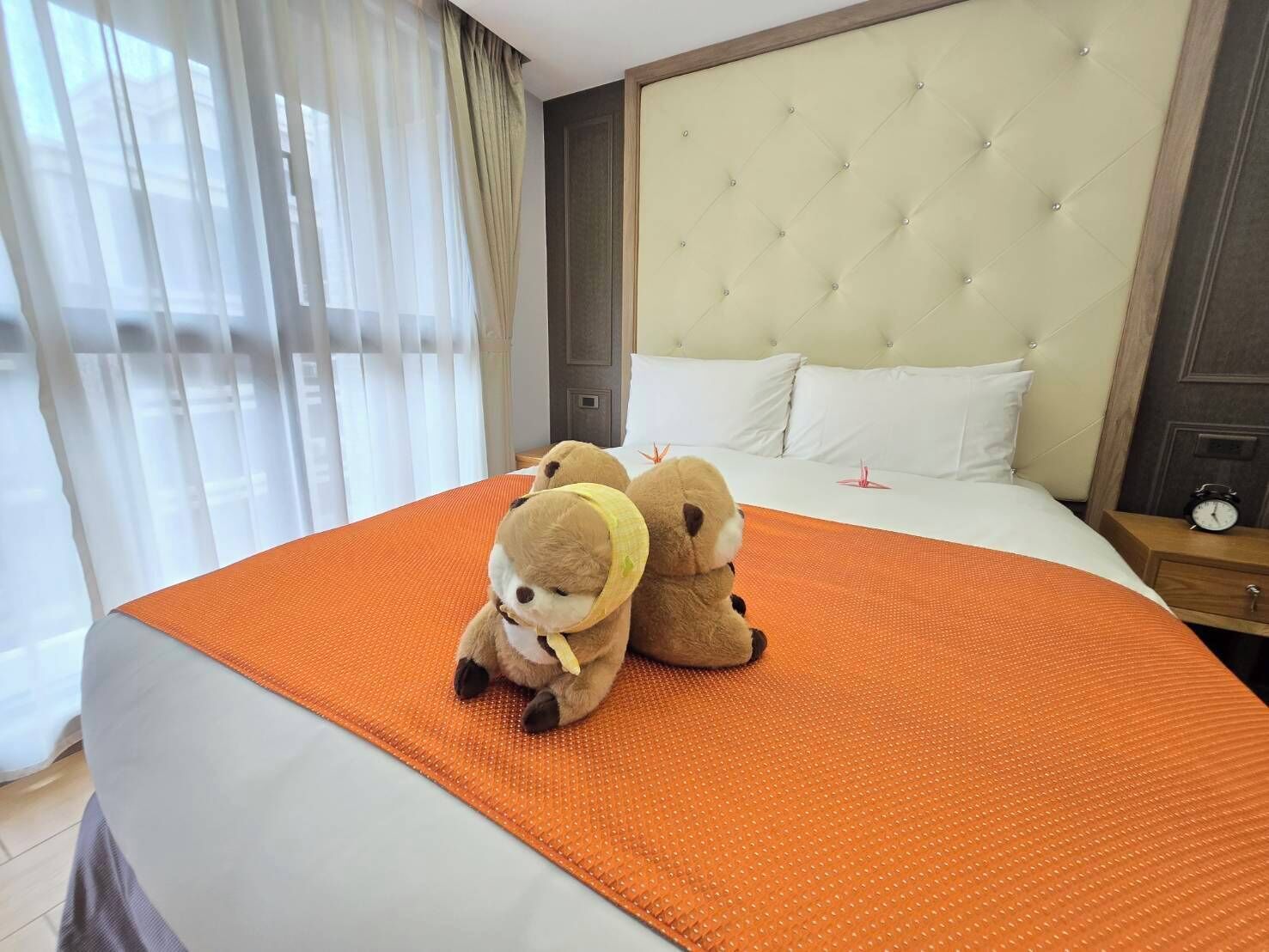 A hotel room with a large bed, a tufted headboard, and two plush toys on the bed.