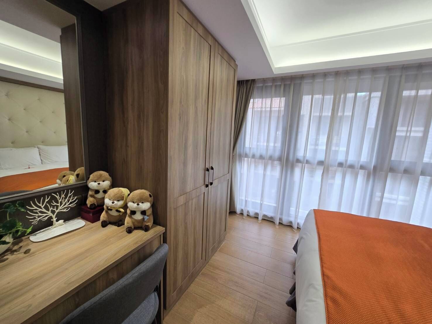 Une chambre d’hôtel avec un lit, une armoire en bois, un miroir et une fenêtre avec des rideaux légers.