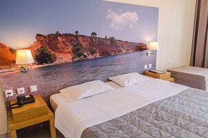 Habitación de hotel con cama, dos lámparas de noche, mesita de noche con teléfono y un mural de paisaje costero.