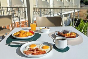 A breakfast table with croissants, eggs, bacon, toast, and a cup of coffee on a balcony with metal railings and chairs. - Nereides Hotel Apartments (Alonissos)