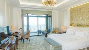 Quarto de hotel com uma cama grande, uma escrivaninha, uma cadeira e um candelabro. Há uma janela grande com vista para o exterior.
