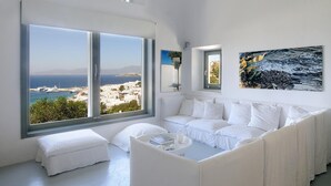 A modern, white-themed living room with large windows offering a view of the sea and a white building. - Great location over Mykonos Town, 294sqm 5 Bedrooms amazing sunset & sea views  (Mikonos)