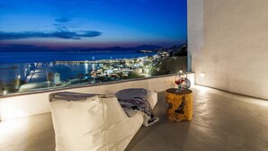 A balcony with a view of a coastal town at dusk, a cushioned seating area, and a small wooden table with a glass of red wine. - Great location over Mykonos Town, 294sqm 5 Bedrooms amazing sunset & sea views  (Mikonos)