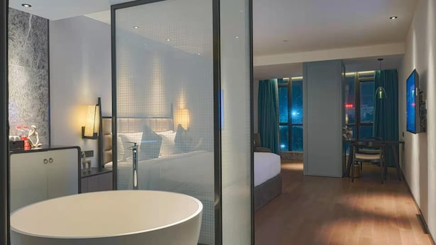 A modern hotel room with a large bathtub, a bed with a view of the city, a dining area, and a separate bathroom. - Paco Hotel (Shenzhen)