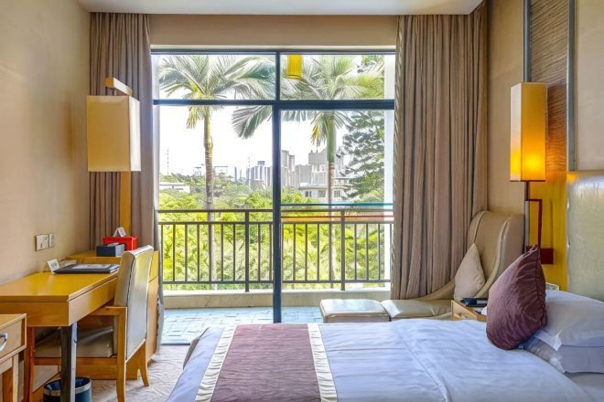 Une chambre d’hôtel équipée d’un lit, d’un bureau, d’une chaise et d’un balcon offrant une vue sur les palmiers et les bâtiments.