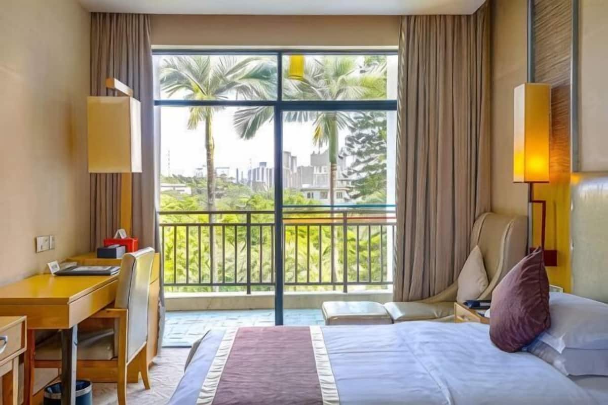 A hotel room with a bed, desk, chair, and a balcony with a view of palm trees and buildings.