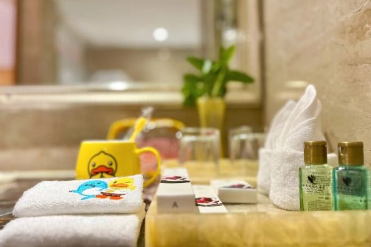 Une salle de bain avec un porte-serviettes sur lequel est posée une serviette ornée de motifs de canards, une tasse jaune décorée d’un canard et deux flacons de lotion.