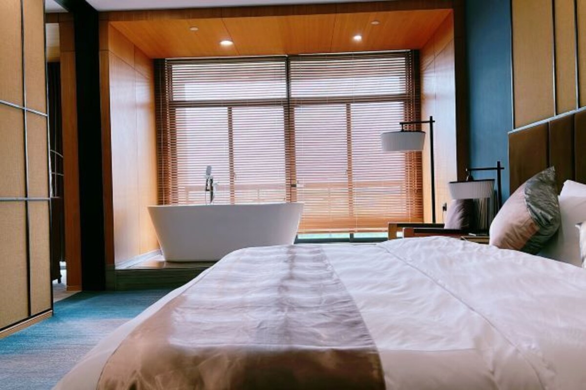 A modern hotel room with a large bed, a bathtub, and a view through floor-to-ceiling windows.
