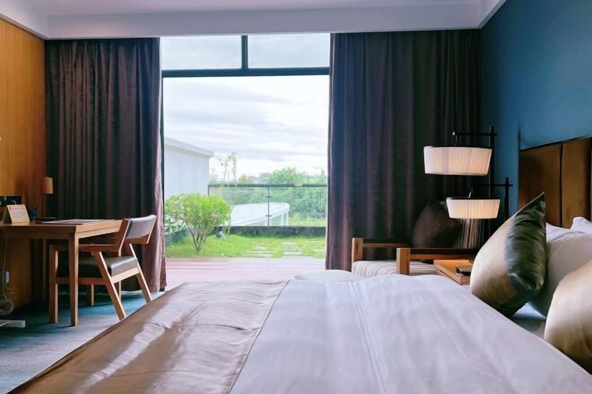 A hotel room with a large bed, a desk, and a chair. There is a view of greenery outside the window.