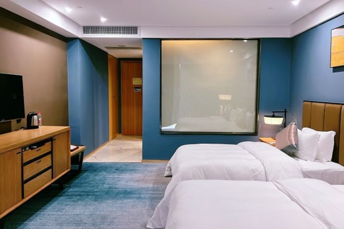 A hotel room with two beds, a TV, a desk with a lamp, and a large window.