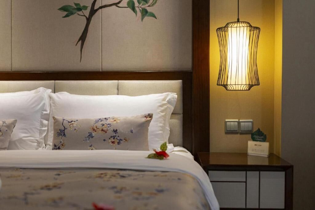 A hotel room with a bed, a nightstand, a wall-mounted lamp, and a painting of a tree branch.