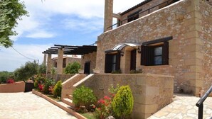 Un edificio de piedra con un espacio exterior techado, escalones que conducen a una puerta y un jardín con diversas plantas.