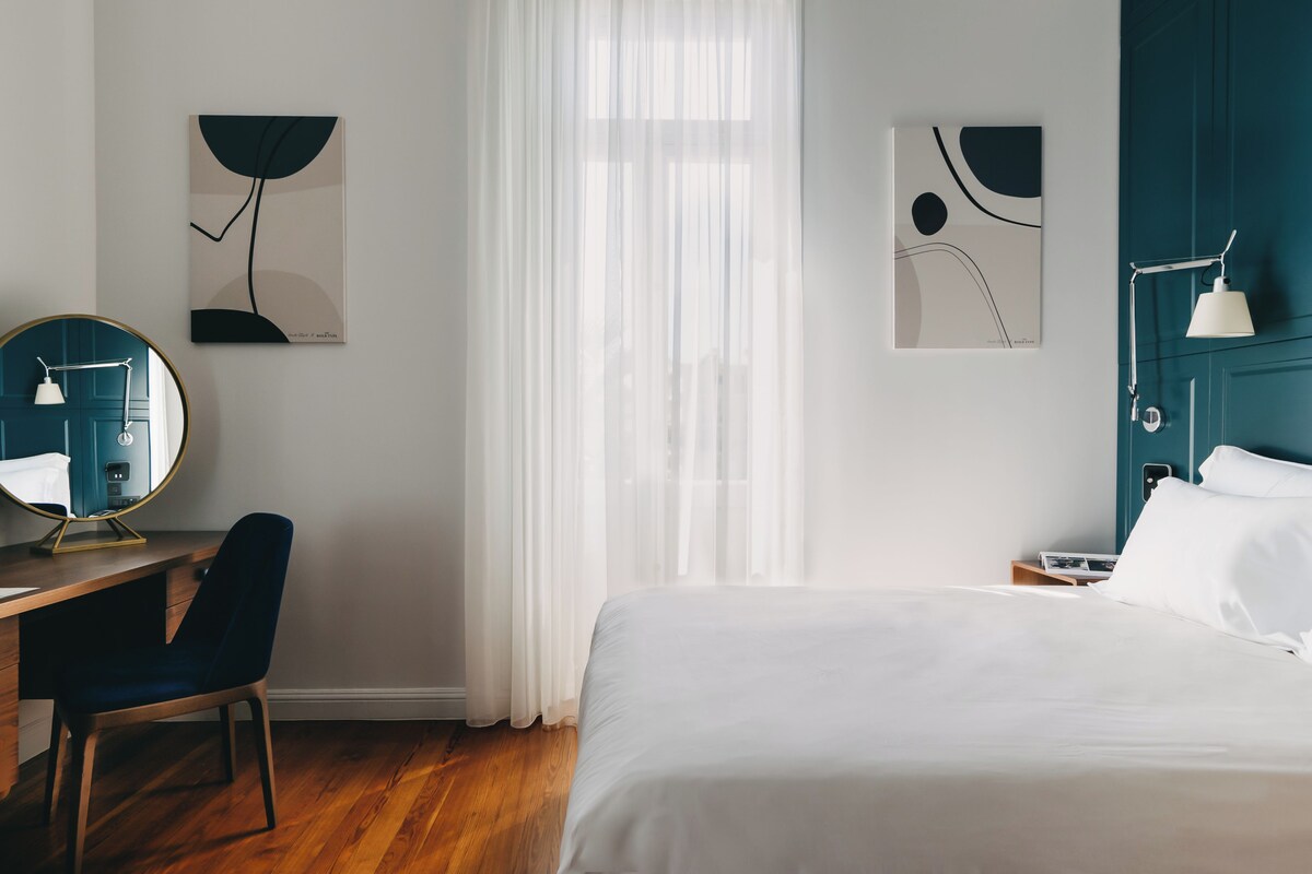 A hotel room with a bed, a desk with a chair, a round mirror, and two wall-mounted artworks.