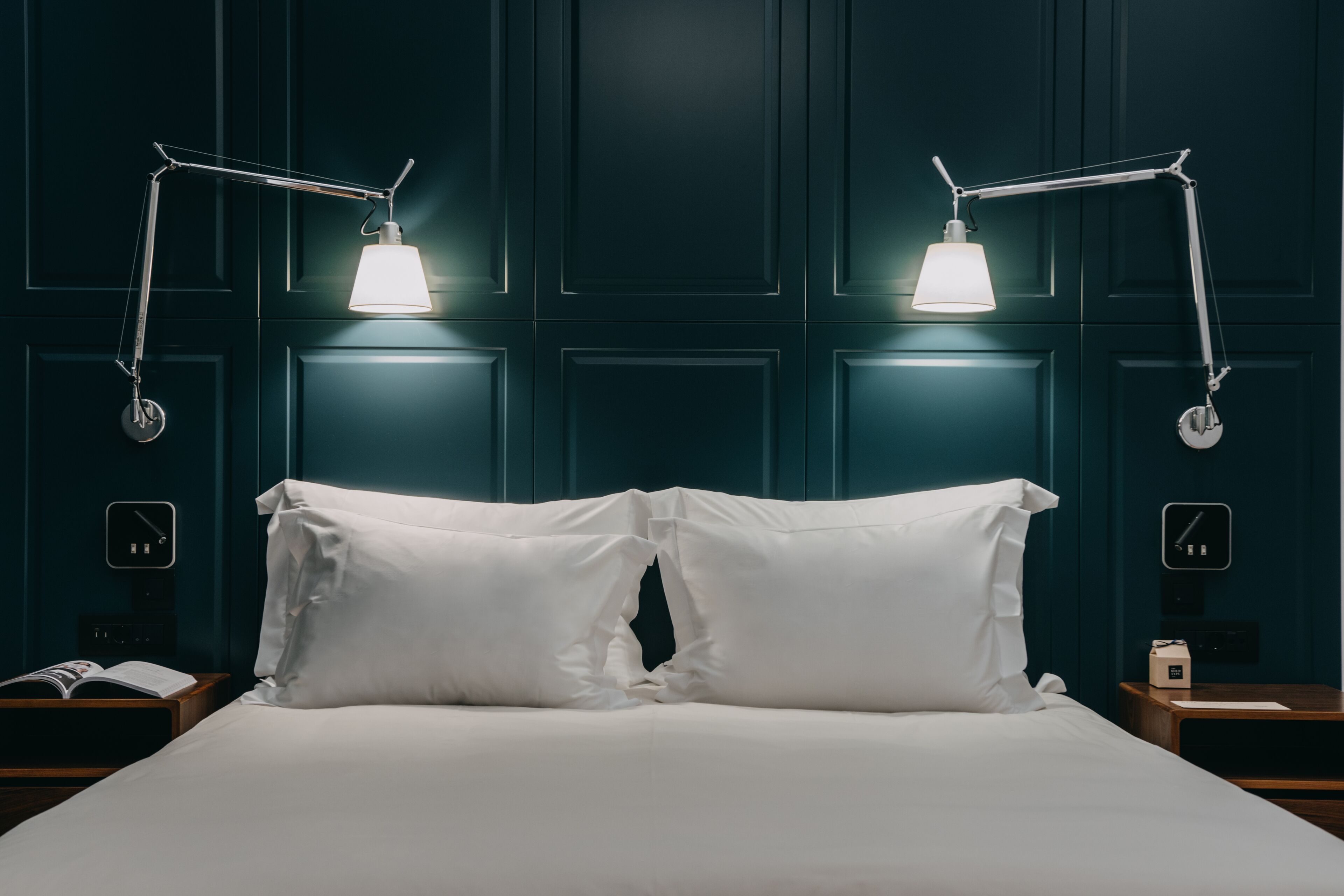 A neatly made bed with white linens, two bedside tables with clocks, and two wall-mounted lamps.