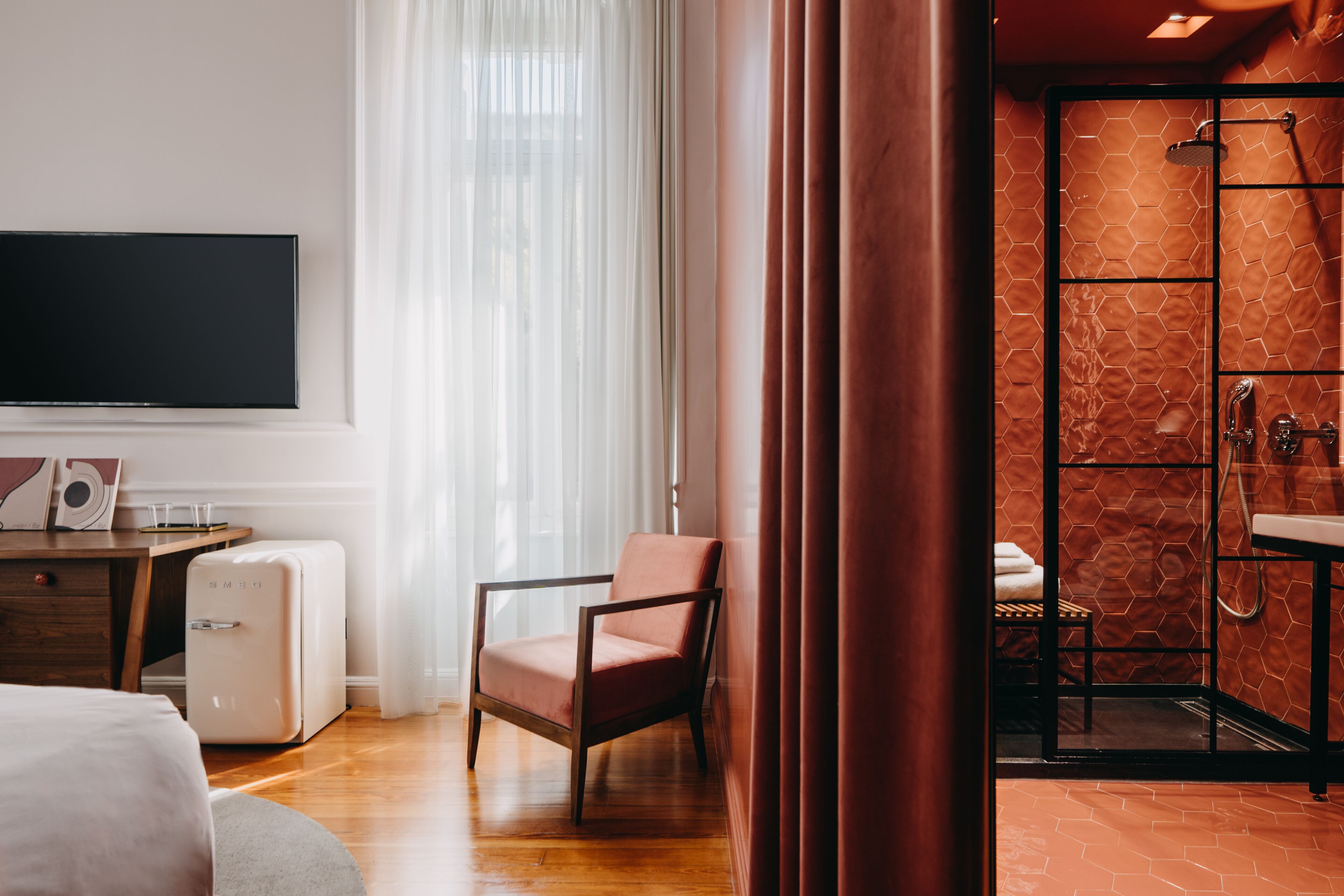 Una camera d'hotel moderna con TV a schermo piatto, una scrivania in legno, un frigorifero in stile vintage, una poltrona comoda e un box doccia con piastrelle ad esagono.