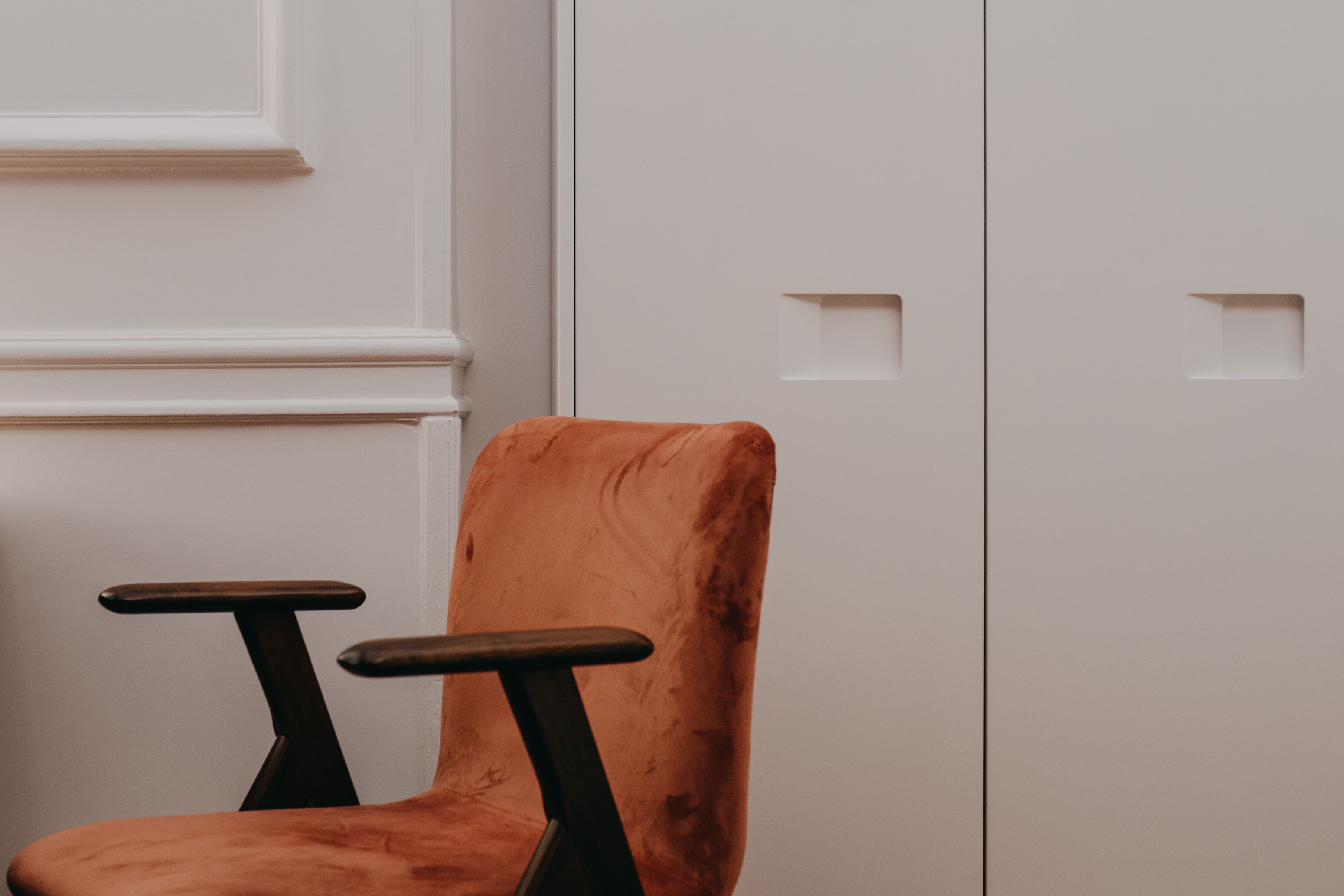 Una silla con respaldo alto y reposabrazos, ubicada frente a una pared blanca con molduras decorativas y una puerta con manija.