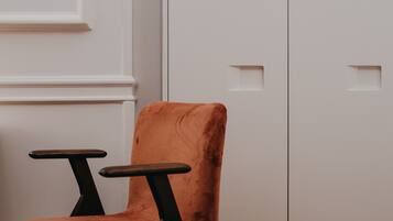 Una silla con respaldo alto y reposabrazos, ubicada frente a una pared blanca con molduras decorativas y una puerta con manija.