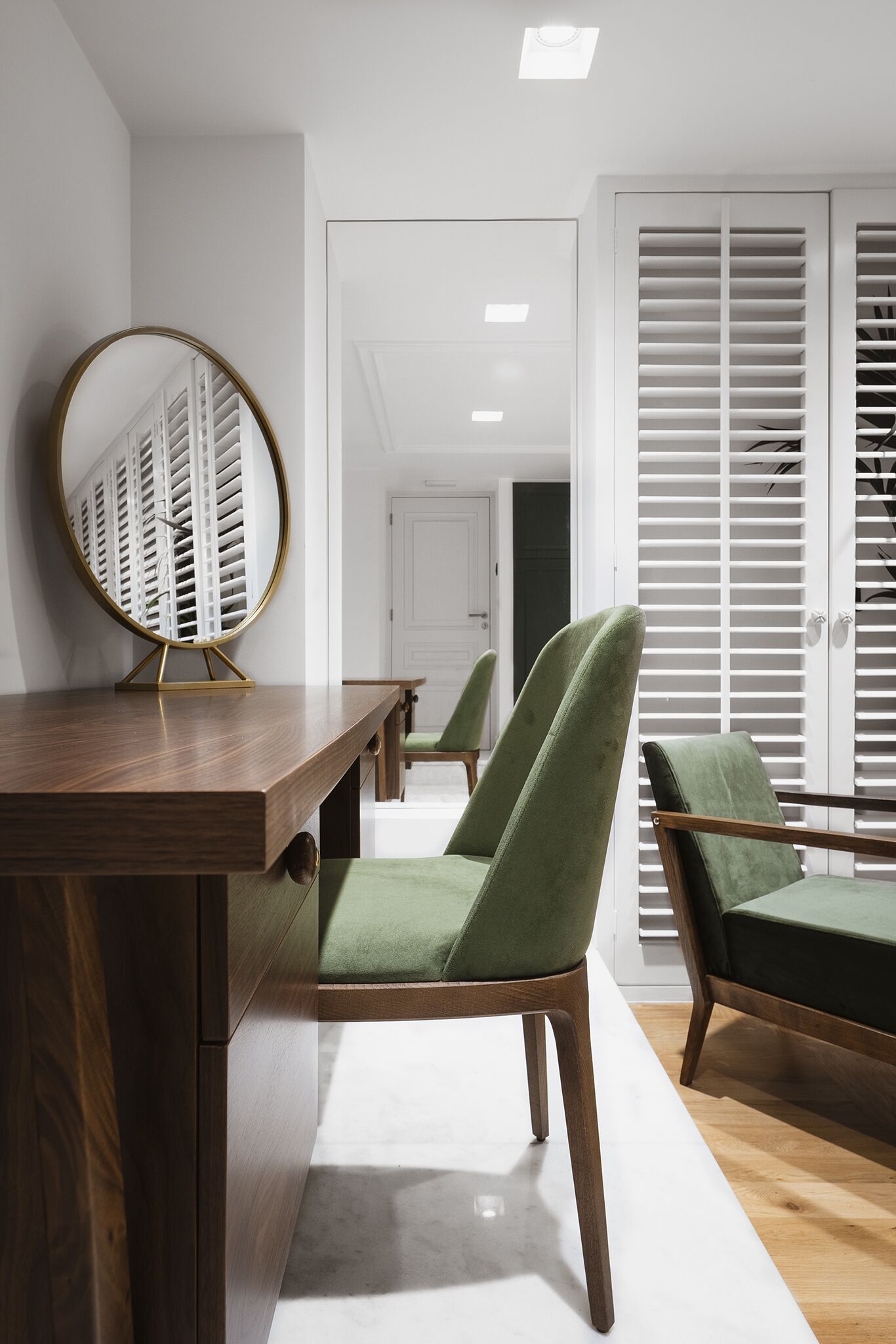 A modern room with a wooden desk, green upholstered chairs, a round mirror, and a white closet with sliding doors.
