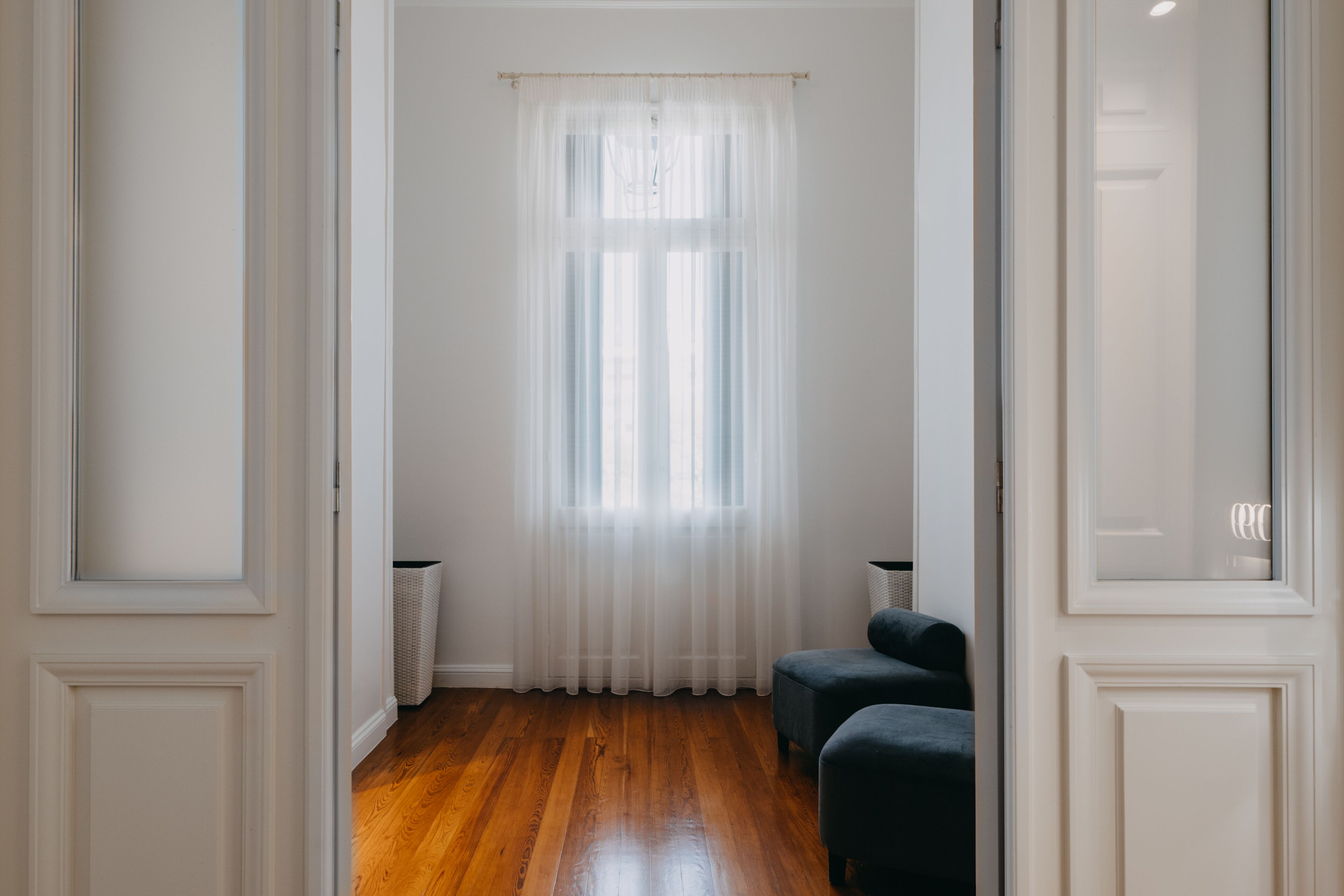 Een kamer met een houten vloer, een raam met transparante gordijnen en twee donkere fauteuils.