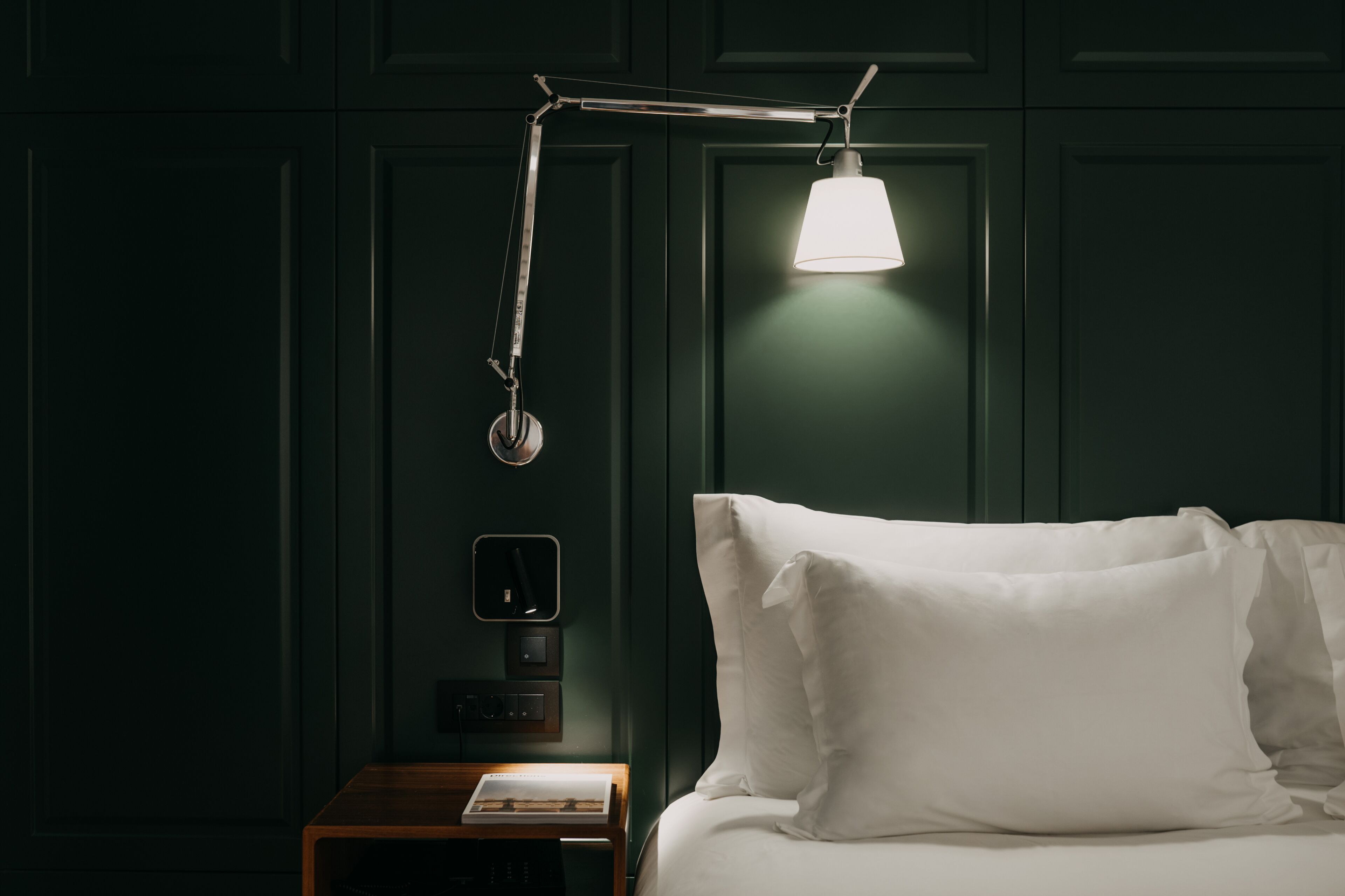 Una cama bien tendida con sábanas blancas, una mesita de noche con un libro, una lámpara de escritorio moderna y un panel de control en la pared.