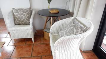 Zwei weiße Korbstühle mit gemusterten Kissen, ein kleiner runder Tisch mit einer gewebten Tischmatte und eine Vase mit einer roten Blume.