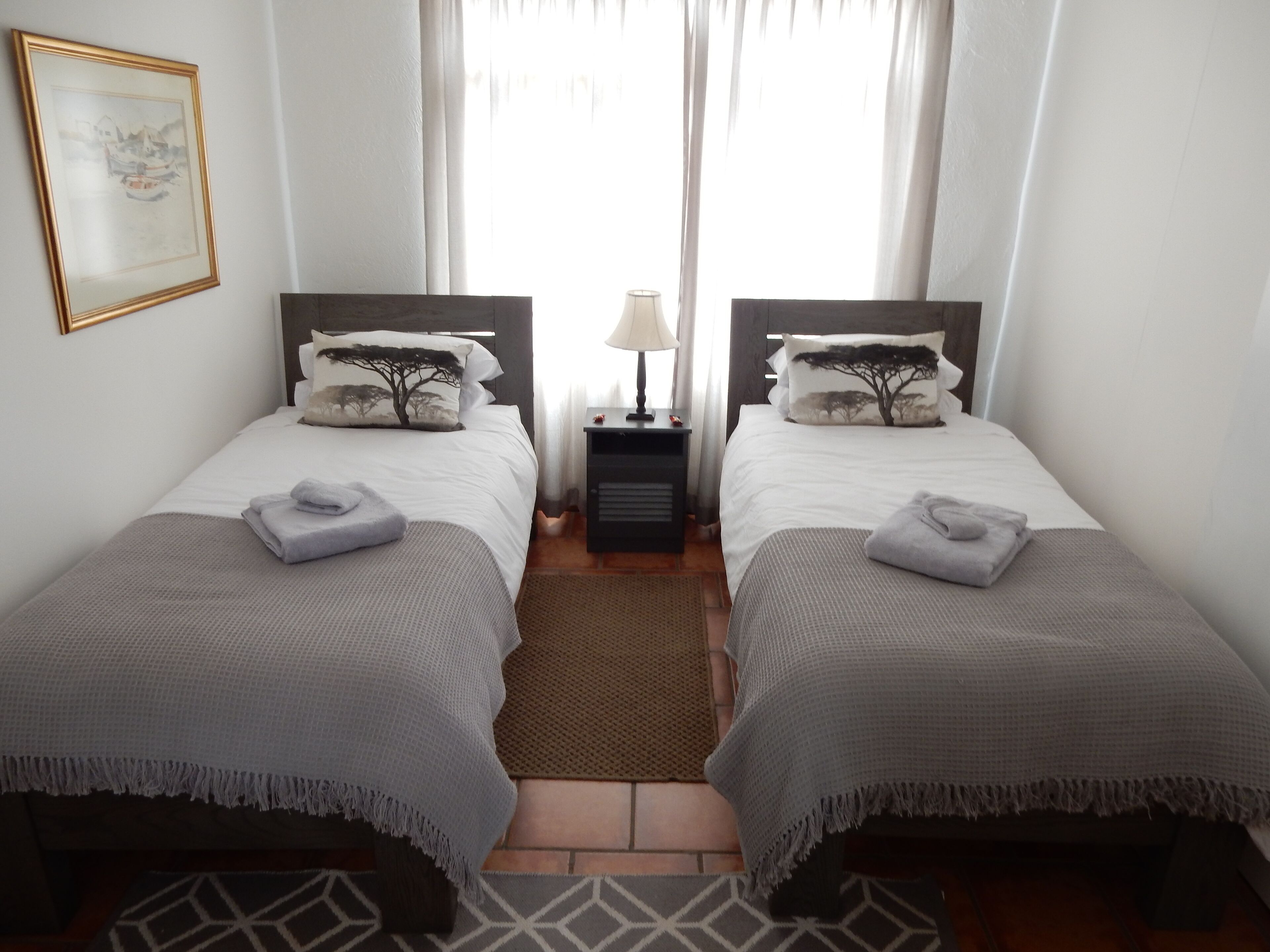 Une chambre d’hôtel avec deux lits, chacun recouvert d’une couverture grise assortie aux oreillers, une petite table de chevet avec une lampe et un tableau encadré au mur.