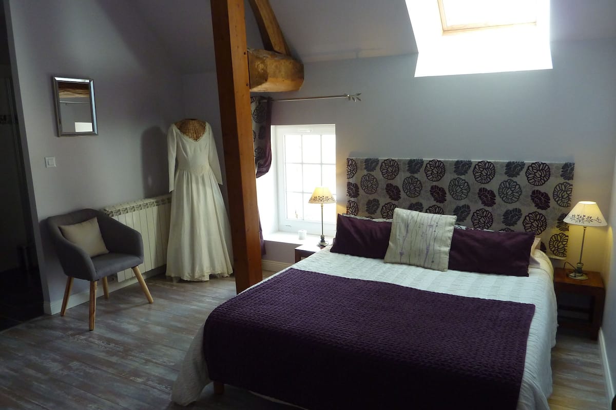 A bedroom with a large bed, a chair, a dress on a mannequin, and a window with natural light.