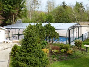 Un invernadero con techo transparente, una piscina y un jardín con diversas plantas y un banco.