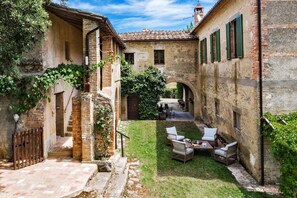 Un patio con zona verde, mobiliario exterior y un edificio de piedra con tejado de baldosas.