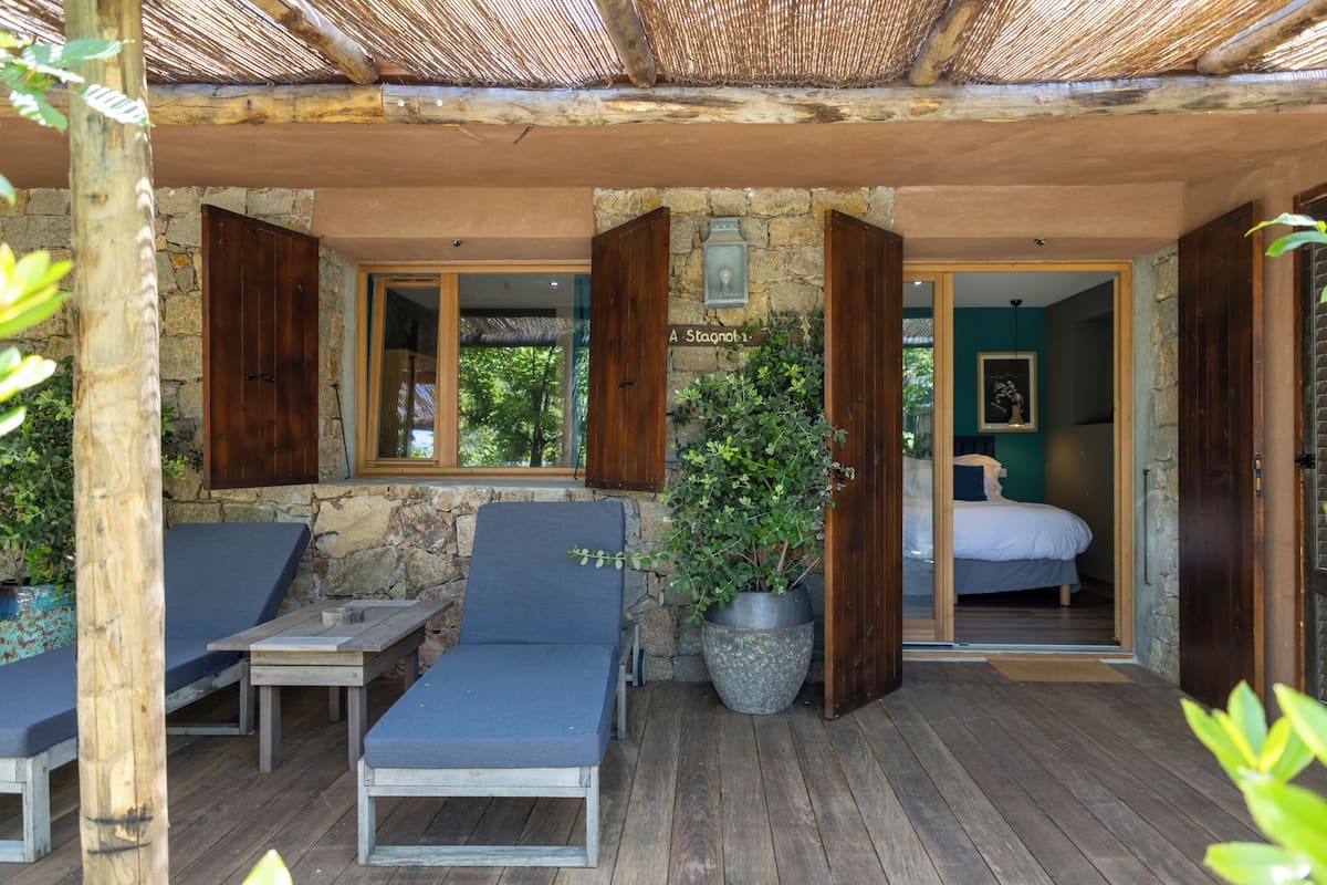 A covered outdoor seating area with lounge chairs, a stone wall, and a potted plant.