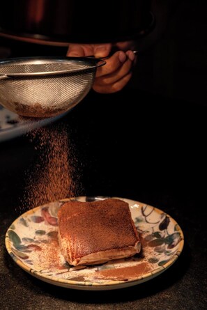 A person holding a strainer over a plate with a food item, possibly a dessert, on a patterned plate. - Villa Kirssis (Pietrosella)