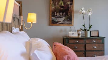 A neatly made bed with pillows, a bedside lamp, a painting, a dresser with drawers, a vase with flowers, and a framed picture.
