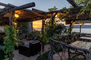 A rustic outdoor seating area with wooden tables and benches, surrounded by greenery and overlooking a body of water. - Villa Kirssis (Pietrosella)