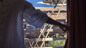 Una persona apoyada en una estructura de madera junto a una ventana que da a una vista costera con una montaña al fondo.