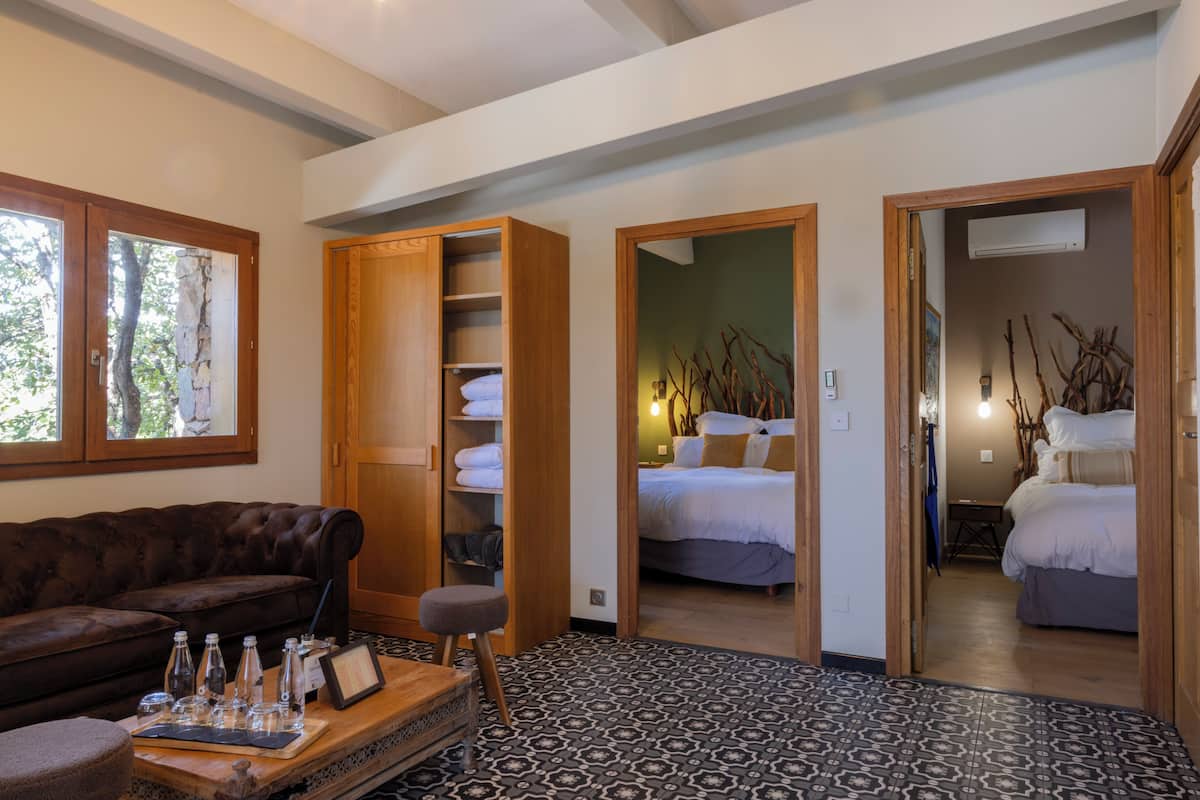 A room with a brown leather sofa, a wooden side table with glasses, a patterned floor, a bed with white bedding, and a wooden wardrobe.