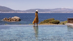 Una piscina cristalina con una persona de vestido amarillo y sombrero blanco, un afloramiento rocoso y un paisaje urbano distante.